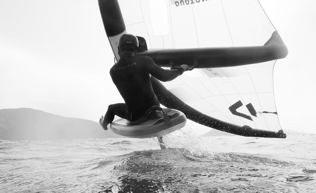 Ein Wingfoiler der sitzend auf dem Board und Foil über das Wasser schwebt - moody Actionfoto in schwarz-weiß mit hoher Belichtung für maavr (maavr-foil)