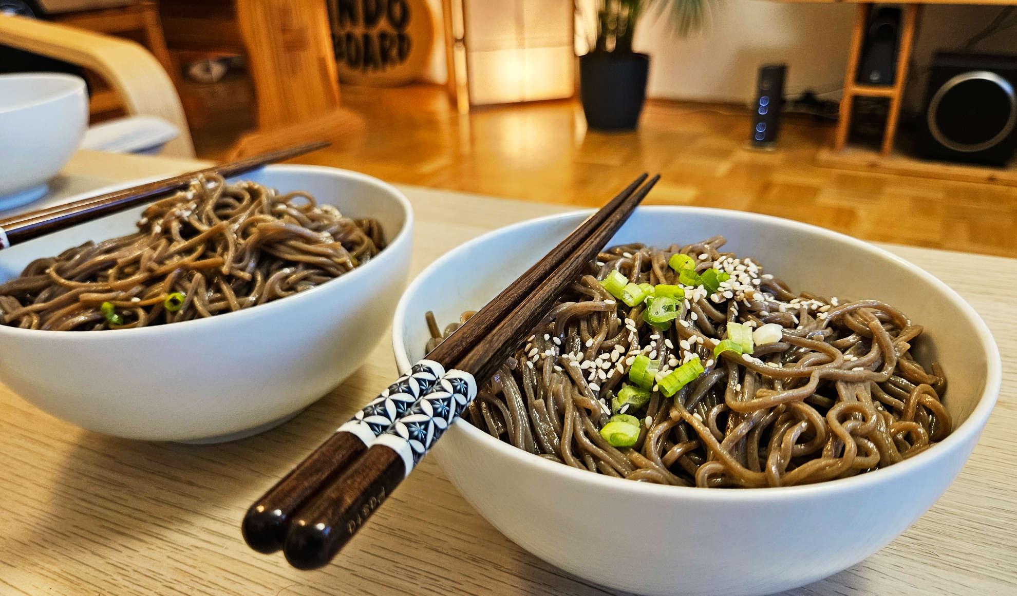 Soba mit Frühlingszwiebeln und Sesam als Topping in einer Bowl auf einem Tisch, mit indirekter Beleuchtung im Hintergrund - Coverfoto eines Rezept-Tipps in der Kategrorie Foiler's Kitchen bei maavr