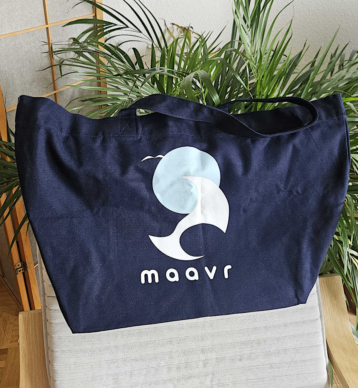 Große dnkelblaue Strandtasche aus Baumwolle mit maavr Logo auf Lounge-Stuhl vor einer Palme 