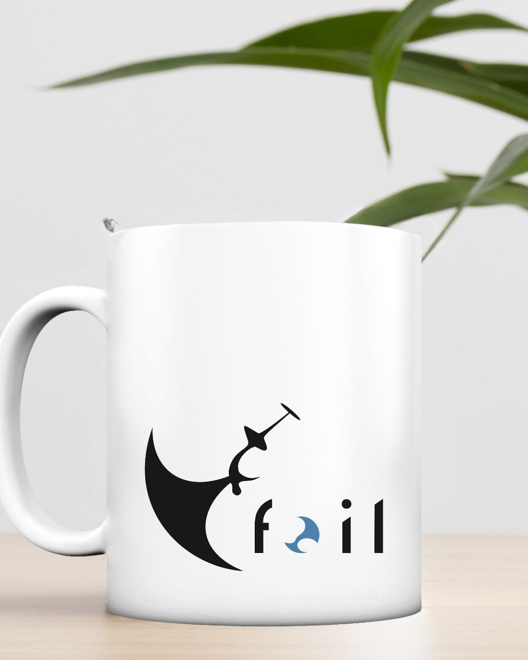 Weiße Matte Keramiktasse  mit Wingfoil-Motiv und "Foil"-Slogan auf einem Tisch mit einer Pflanze im Hintergrund