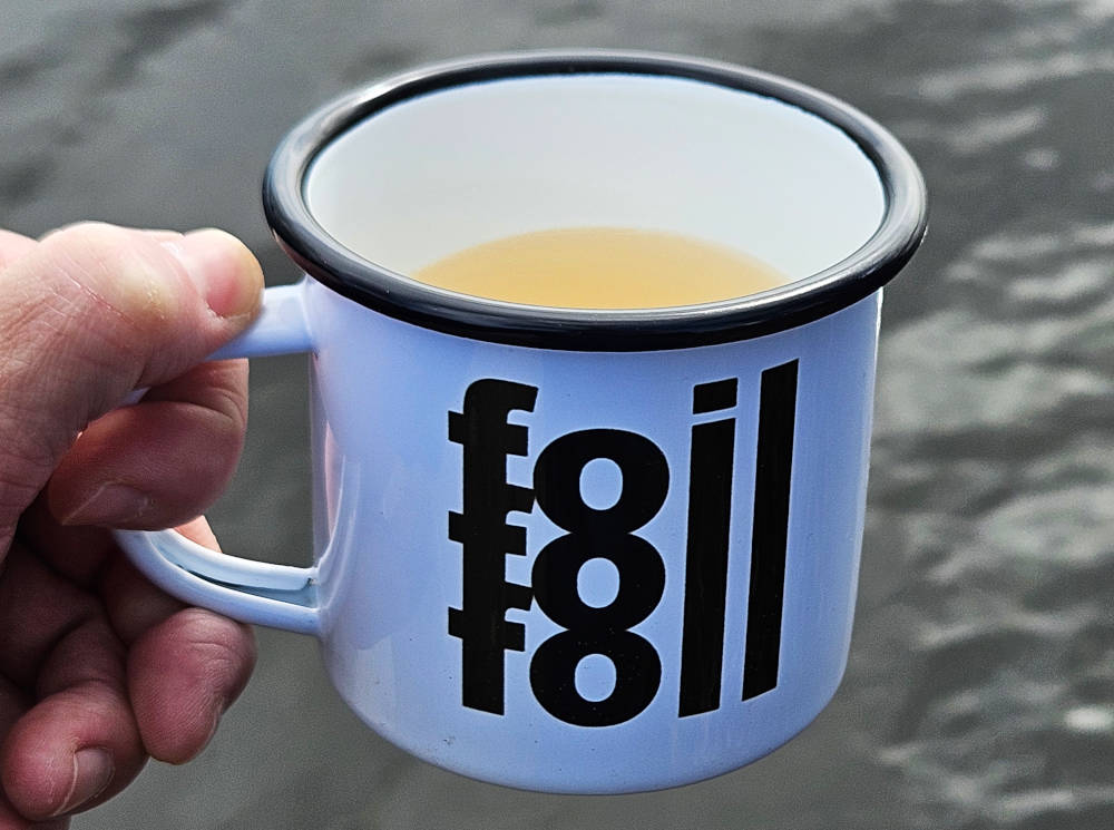 Hand hält mit Tee befüllte Emaille-Tasse mit schwarzem Rand und  "Foil Foil Foil"  Schriftzug über Wasser