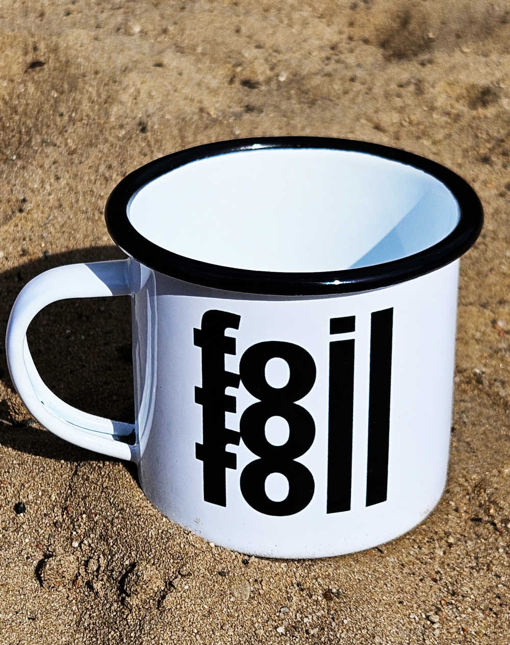 Emaille-Tasse mit schwarzem Rand und schwarzem "Foil Foil Foil" Schriftzug im Sand an einem sonnigen Strand