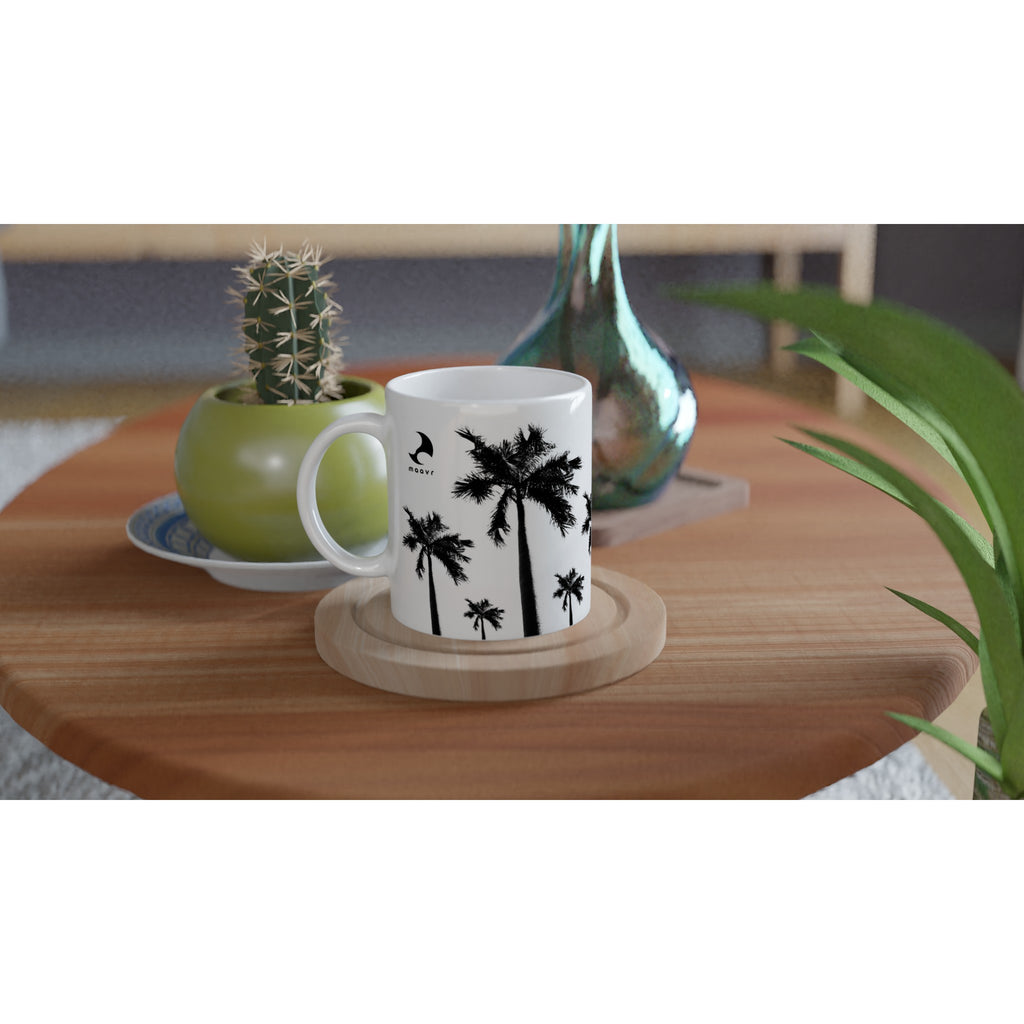 Weiße Keramiktasse mit Palmen-Motiven und  maavr Logo auf Holztische arrangiert mit Kaktus und Vase