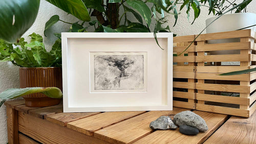 Druck einer Kaltnadelradierung mit Wingfoil-Motiv in weißem Rahmen auf Holzkiste mit Pflanzen und Holzkisten arrangiert