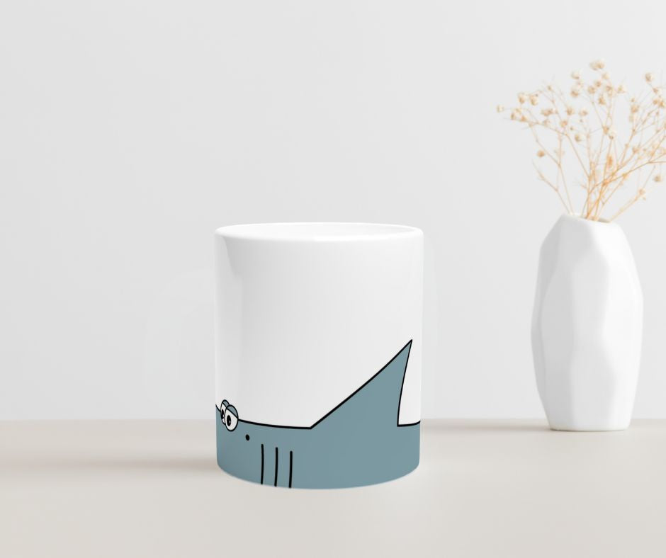 Weiße Keramiktasse mit blauem Cartoon-Hai-Motiv im unteren Drittel der Tasse  auf  einem Tisch mit einer Vase