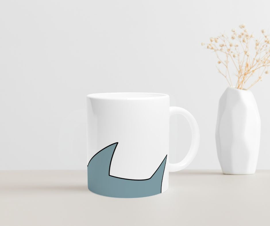 Weiße Keramiktasse mit blauem Cartoon-Hai-Motiv im unteren Drittel der Tasse  auf  einem Tisch mit einer Vase