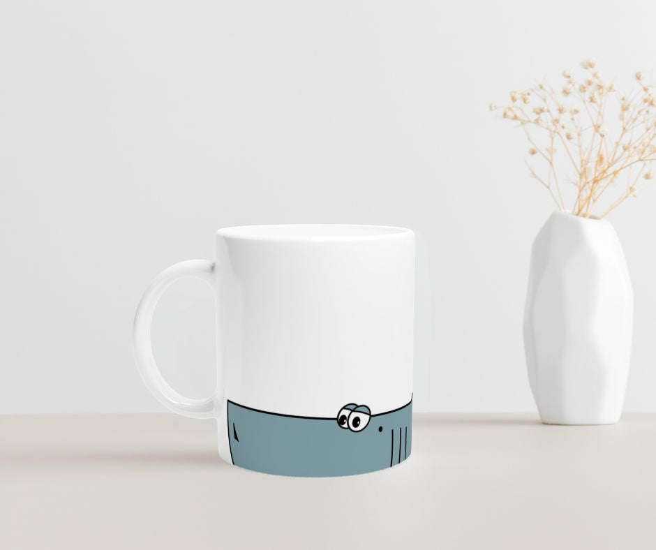 Weiße Keramiktasse mit blauem Cartoon-Hai-Motiv im unteren Drittel der Tasse  auf  einem Tisch mit einer Vase