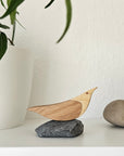 Holzvogel aus Eiche und Birke auf Naturstein, dekoriert auf weißem Regal neben Pflanze und Kieselsteinen