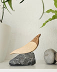 Holzvogel aus Eiche und Birke auf dunklem Naturstein, arrangiert auf weißem Regal neben Pflanzentöpfen