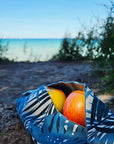 Snacktasche mit blauem Pflanzenmuster, gefüllt mit Äpfel, die herausragen - auf einem Baumstamm mit Küstenhintergund