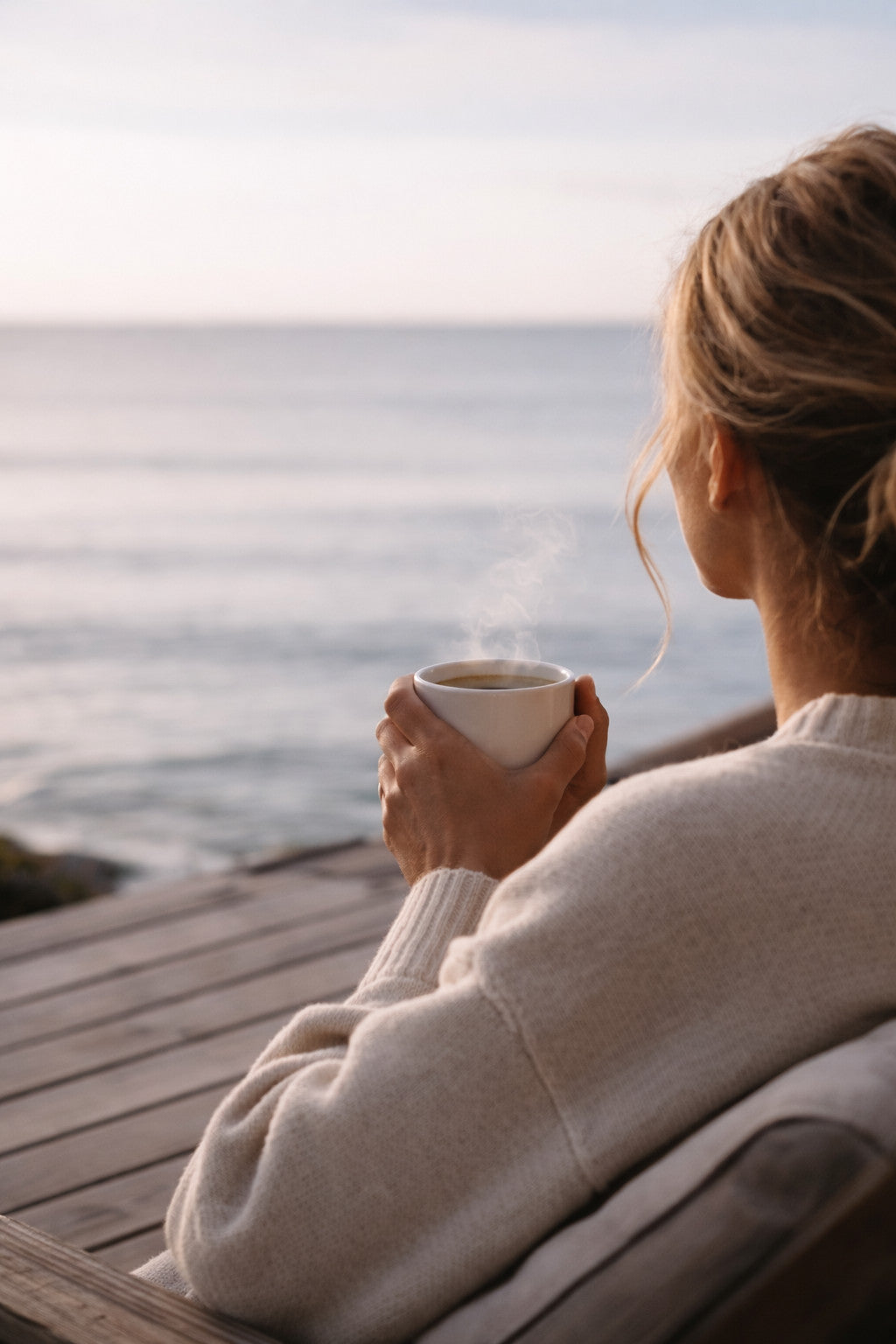 Frau mit Keramiktasse in der Hand sitzt auf Bank und schaut aufs Meer