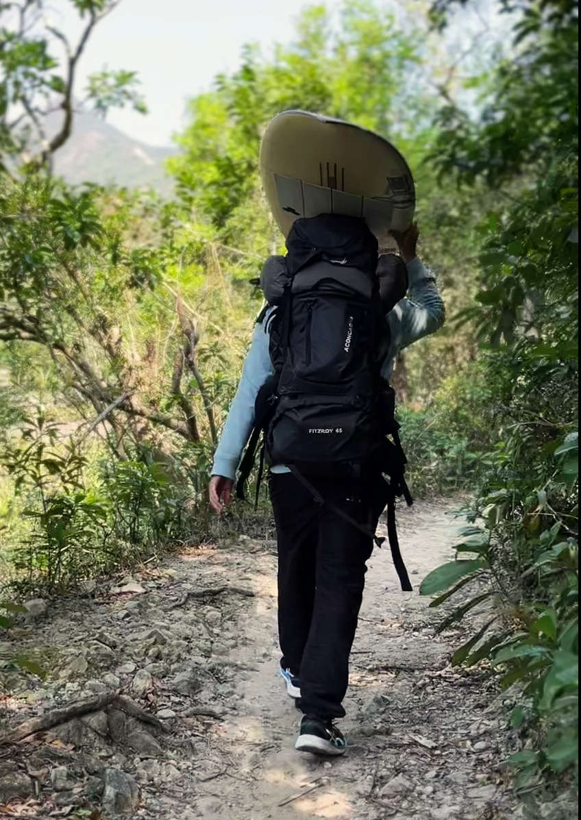 Person mit Surfboard und Rucksack auf Wanderweg in der Natur auf dem Weg zum Meer