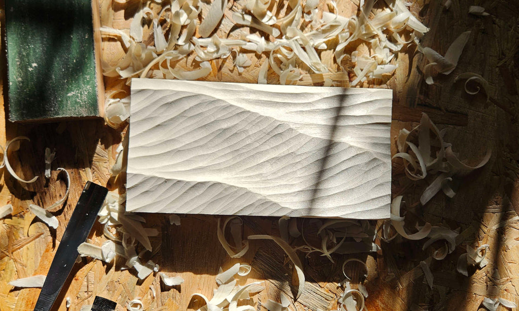 Gerahmtes Wellenrelief aus hellem Holz, neben Holzspänen und Schnitzutensilien auf eine Holzplatte im Sonnenschein in einer Werkstatt