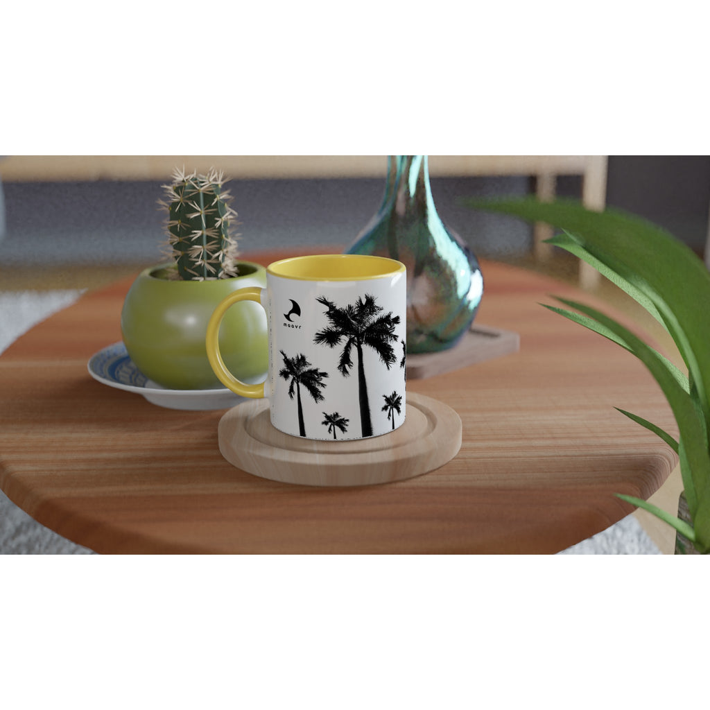 Weiße Keramiktasse mit Palmen-Motiven, maavr Logo, gelber Innenfläche und gelbem Henkel auf Holztisch arrangiert mit Kaktus und Vase