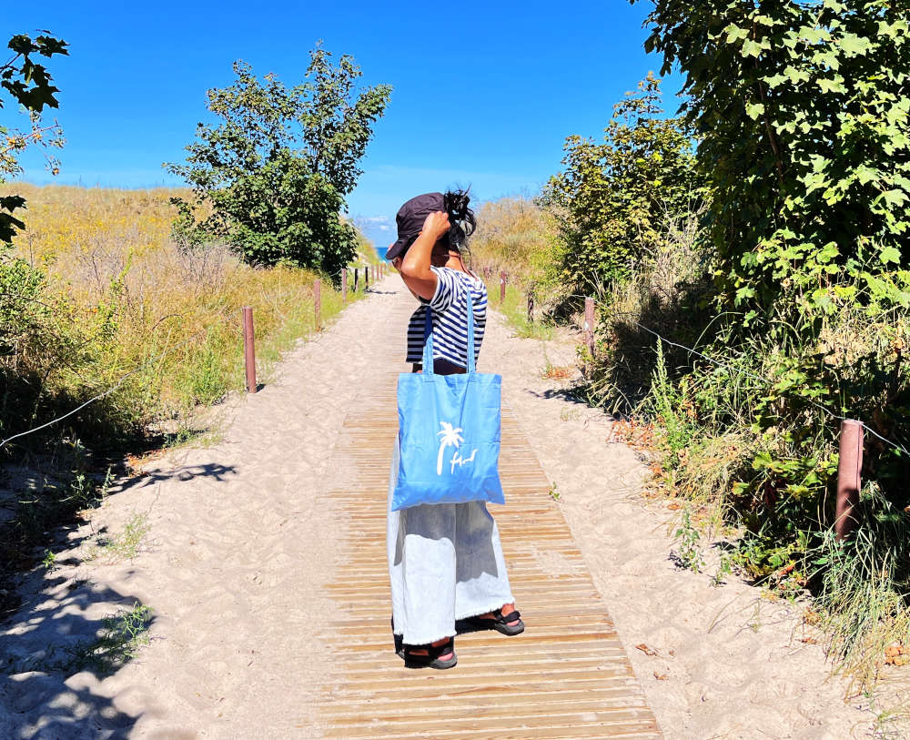 Blaue Tote Bag um die Schulter hängend getragen auf einem Holzweg zum Meer von einer Frau getragen, die ihr Cappie richtet  - sommerliches Produktbild von maavr