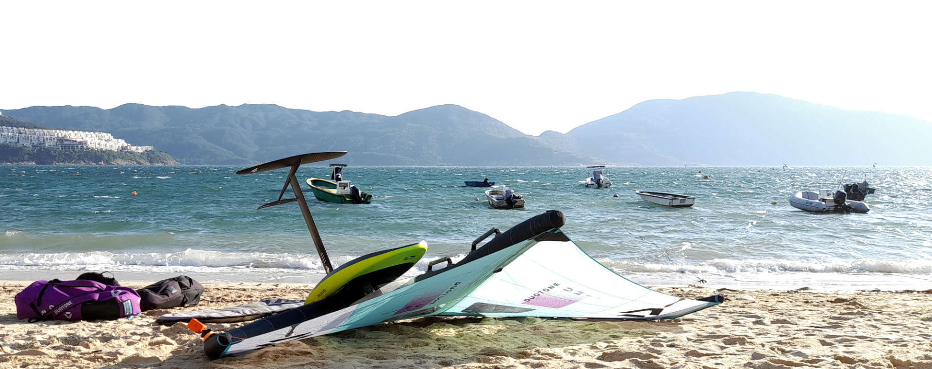 Wingfoilequipment im Sonnenschein am Strand einer Meeresbucht in Asien, umrandet von Bergen - Verbindung von Action auf dem windigen Wasser und Entspannung am Strand - Lifestylefoto für maavr (maavr-foil)