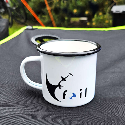 Weiße Emaille-Tasse mitschwarzem Rand, Wingfoil-Backflip-Motiv und "Foil"-Schriftzug auf Campingtisch