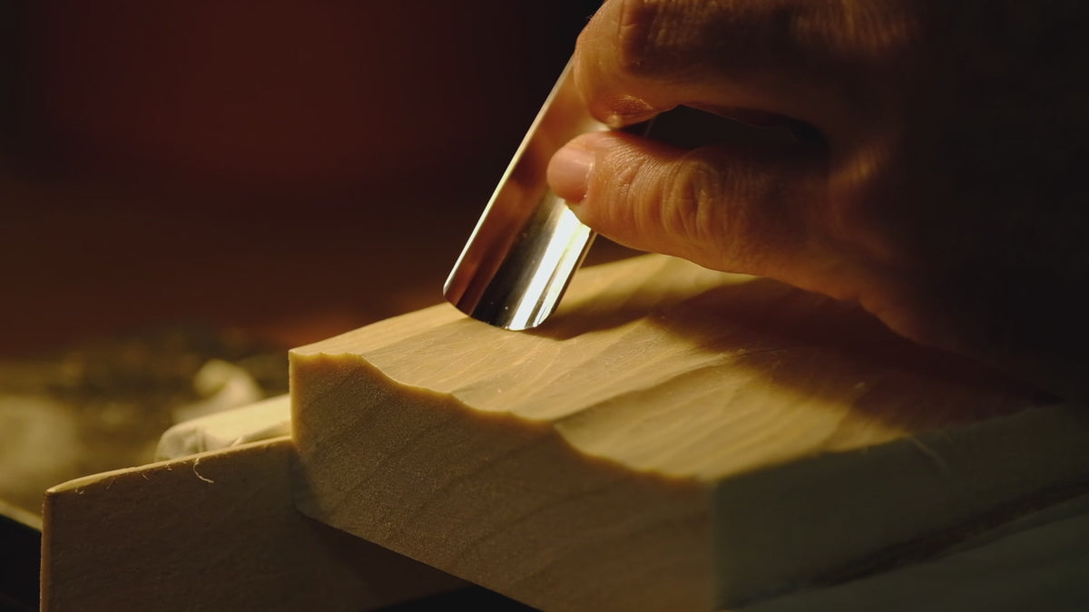 Video mit Schnitzszene und in Holz geschnitztes Wellenrelief auf Werkstatttisch neben Schnitzeisen und Holzspänen im Sonnenlicht